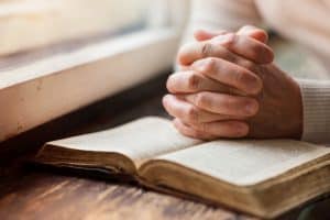 Woman by window with hands praying over a Bible